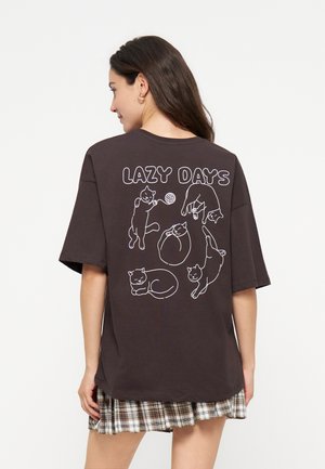 Femme portant un t-shirt sombre oversize avec « LAZY DAYS » et des dessins linéaires blancs de cinq chats joueurs dans le dos, associé à une jupe à carreaux.