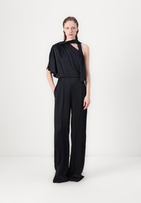 Macacão de cetim preto com um design drapeado de um ombro, apresentando pernas largas e um detalhe de gola segurada. Textura suave, estilo minimalista.