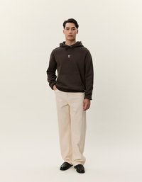 Felpa marrone con cappuccio e cordino, tasca frontale e logo bianco. Abbinata a pantaloni larghi color crema e scarpe nere. Design semplice e casual.