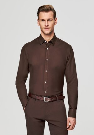 Mężczyzna noszący dopasowaną ciemnobrązową koszulę na guziki i pasujące spodnie z brązowym skórzanym paskiem, stojący na jednolitym jasnym tle.
