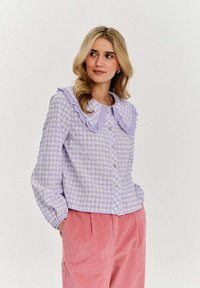 Jasnofioletowa koszula w kratkę z guzikami, z falbaniastym kołnierzykiem i długimi rękawami, w zestawieniu z różowymi spodniami z kordurowej tkaniny. Gładka tekstura materiału.