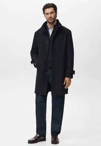 Abrigo de lana azul marino con cuello alto y cierre de botones, combinado con un suéter oscuro y pantalones de mezclilla. Textura suave y diseño clásico.