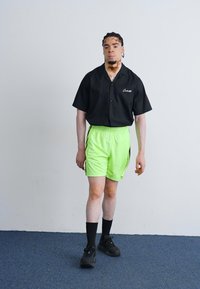 Black short-sleeve button-up shirt, neon green shorts, black socks, and shoes. Smooth fabric with a relaxed fit. Simple design and vibrant color contrast.