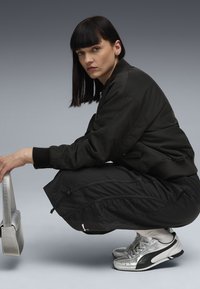 Veste bomber noire avec des poignets côtelés, pantalon cropped noir, sac métallique et baskets argentées. Cheveux lisses et foncés. Arrière-plan neutre.
