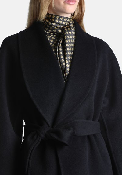 Femme portant un manteau noir ceinturé par-dessus une écharpe en soie à motifs nouée autour du cou, avec de longs cheveux blonds visibles.