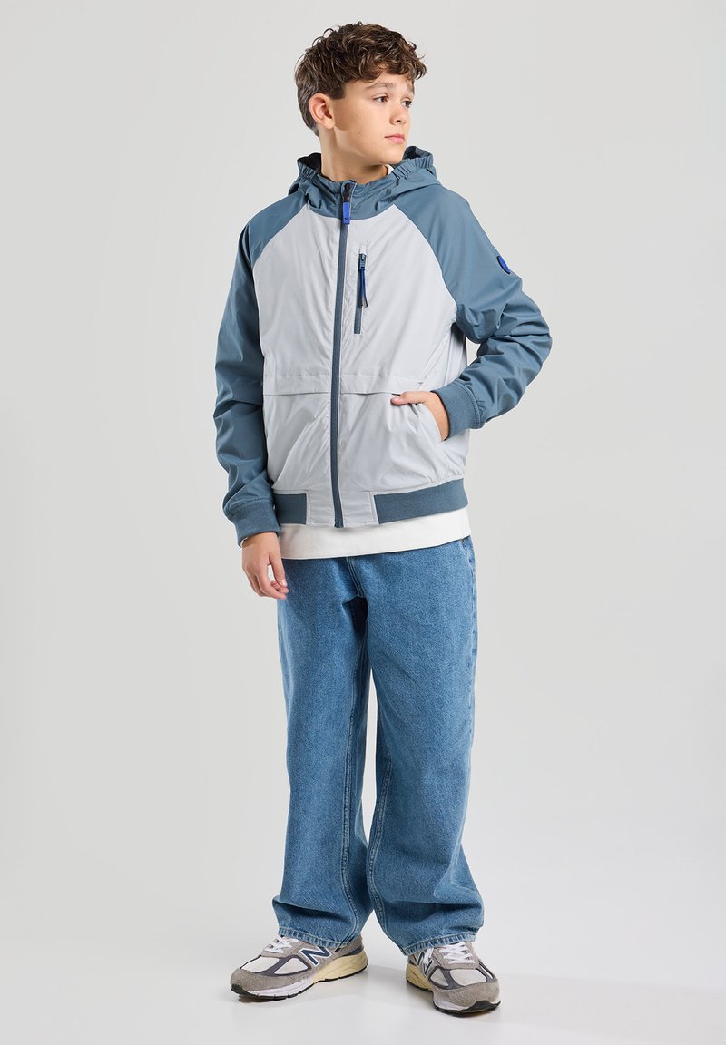 Vaaleanharmaa ja sininen takki, jossa on vetoketjullinen tasku, huppu ja erottuvat hartiapaneelit. Yhdistetty tilaviin sinisiin farkkuihin ja urheilusandaaleihin.