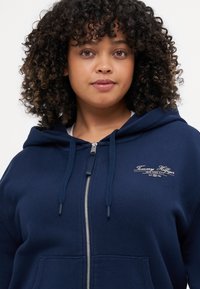 Sudadera con cremallera azul marino, con cordones, material de mezcla de algodón, bolsillo canguro y logo blanco bordado en el pecho. Textura suave.