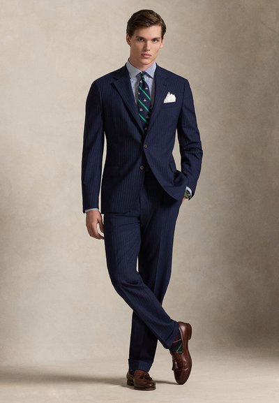 Κοστούμι με ναυτικές ρίγες και εφαρμοστή γραμμή, σε συνδυασμό με γαλάζιο καρό πουκάμισο, γραβάτα με πράσινο μοτίβο και καφέ δερμάτινα παπούτσια.