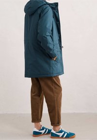 Tyrkysová nepromokavá bunda s kapucí, která má stahovací šňůrku. V kombinaci s hnědými kordovými kalhotami a modrými teniskami s bílými akcenty.