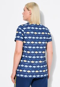 Námořnické tričko s krátkým rukávem, ozdobené opakujícím se potiskem origami lodí v modré, bílé a žluté barvě na pruhovaném pozadí.
