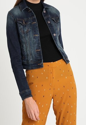 Femme portant une veste en jean foncée sur un top noir et un pantalon jaune moutarde avec de petites lignes blanches et noires, debout devant un fond uni.