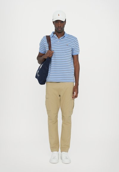 Μπλε ριγέ polo μπλούζα, χακί cargo παντελόνια, λευκά αθλητικά παπούτσια και λευκό καπέλο, κρατώντας μια ναυτική τσάντα με λουράκι στον ώμο.