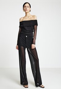 Schwarzes schulterfreies Oberteil mit gerafften Details, kombiniert mit schwarzen, transparenten Weitbein-Hosen mit Knopfakzent. Glatte Textur.