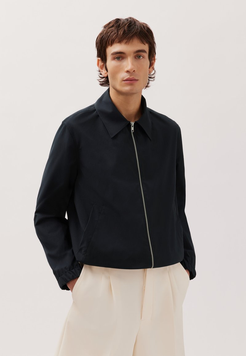 Jeune homme portant une veste noire zippée et un pantalon large beige clair, debout les mains dans les poches contre un fond uni.
