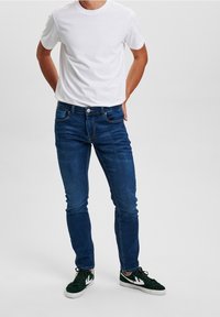 Blå denimjeans med smal passform, femficksdesign och subtil blekning. Kombinerade med en enkel vit t-shirt och mörkgröna sneakers.