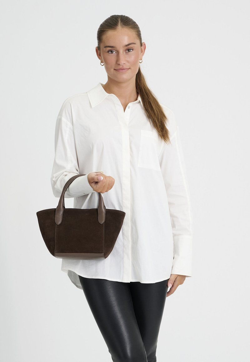 Jeune femme avec une queue de cheval brune portant une chemise blanche et un pantalon noir, tenant un petit sac à main brun foncé sur un fond uni.