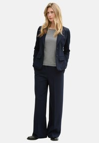 Blazer bleumarin cu două buzunare frontale, asociat cu o bluză cu dungi alb-negru și pantaloni largi bleumarin. Țesătură netedă cu o textură subtilă.