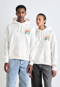 Sudaderas color crema con un bolsillo frontal, que presentan un colorido logo bordado de "Tommy". Una persona lleva jeans azul claro, y la otra jeans oscuros.