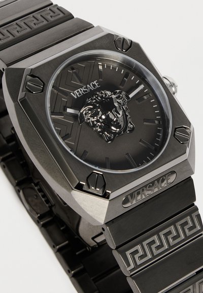 Montre bracelet noire Versace avec emblème tête de Méduse, bracelet en métal à motif géométrique et boîtier angulaire en acier inoxydable avec vis visibles.