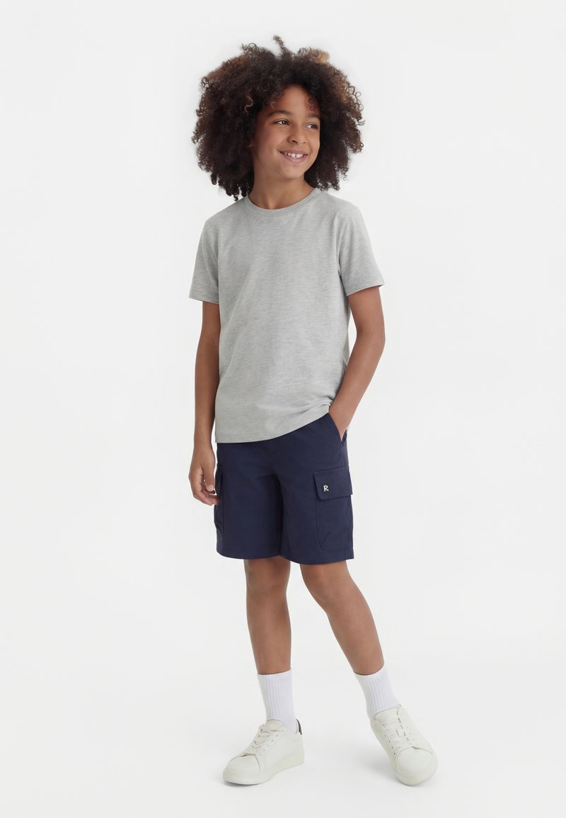 Χαμογελαστό παιδί με σγουρά μαλλιά που φοράει ανοιχτό γκρι κοντομάνικη μπλούζα, σκούρα μπλε cargo σορτς, λευκές κάλτσες και λευκά αθλητικά παπούτσια, στέκεται με το ένα χέρι στην τσέπη.