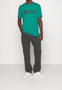 Grøn bomulds t-shirt med teksten "BOSS" i grå skrift, parret med mørkegrå denimjeans og hvide sneakers. Modellen står i en neutral baggrund.