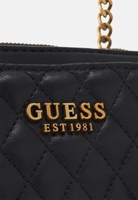 Czarna pikowana torebka z złotym logo "GUESS EST 1981". Materiał wydaje się gładki o błyszczącej fakturze, z metalicznymi detalami.