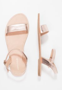 Sandali flat color oro rosa con una cinturino metallico liscio, una soletta beige morbida e una cinghia alla caviglia regolabile. Design minimalista senza decorazioni.
