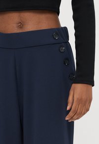 Nærbilde av en person som har på seg marineblå bukser med fire svarte knapper på linningen og en svart langermet kort topp som viser bar mage.