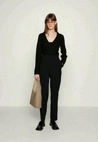 Woman wearing black V-neck sweater, black tapered pants, black loafers, and large beige tote bag, standing against plain light wall.