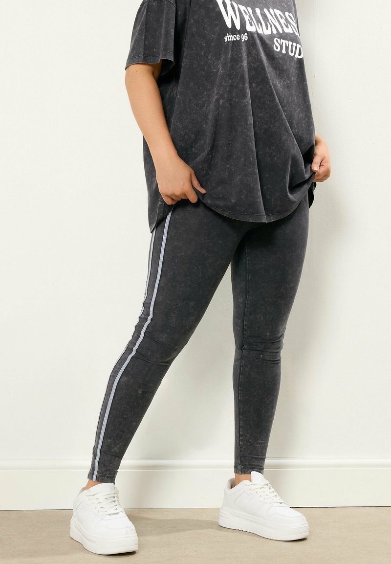 Persoon die zwarte acid-wash legging draagt met witte zijstrepen, een oversized zwart T-shirt met witte tekst, en witte platform sneakers, staande binnen.