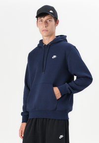 Sweatshirt azul-marinho com um bolso frontal, capuz com cordão e um pequeno logotipo branco da Nike no peito, combinado com calças de desporto pretas.