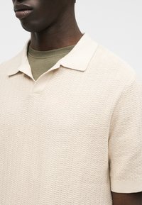 Gros plan sur une personne portant un polo à manches courtes crème texturé par-dessus un t-shirt à col rond vert olive.