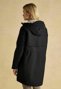 Schwarzer Kapuzenmantel mit weichem, beigem Fleece-Futter, locker sitzend, mit geknöpfter Vorderseite und langen Ärmeln. Rückendetail mit horizontaler Naht.