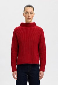 Roter, voluminöser Strickpullover mit hohem, umgeschlagenem Kragen und langen Ärmeln. Hat eine Ripp-Struktur und eine kurze Silhouette. Kombiniert mit dunkelblauen Jeans.