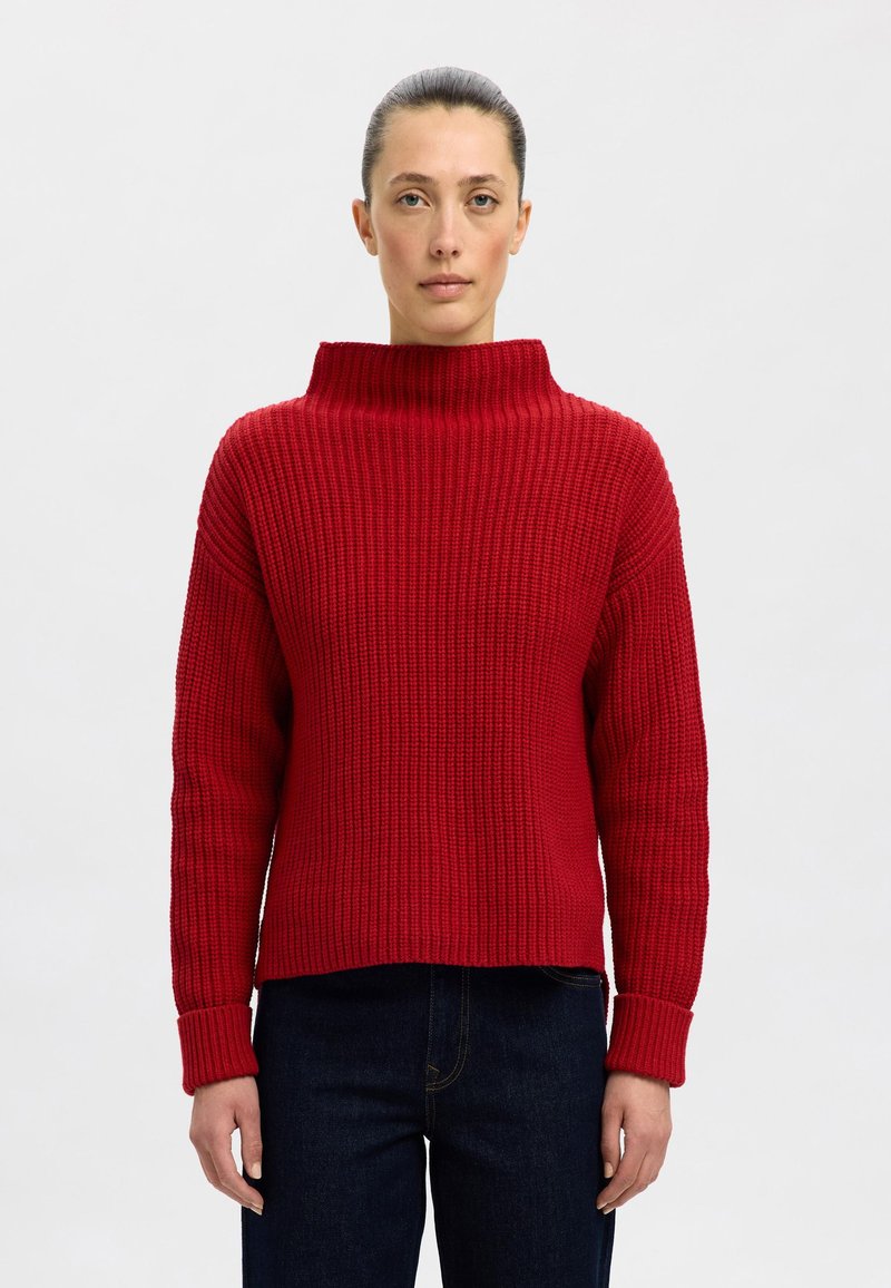 Roter, voluminöser Strickpullover mit hohem, umgeschlagenem Kragen und langen Ärmeln. Hat eine Ripp-Struktur und eine kurze Silhouette. Kombiniert mit dunkelblauen Jeans.