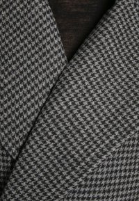 Grijs gestructureerde stof met een houndstooth-patroon, met verschillende tinten grijs en zwart, die een driedimensionaal uiterlijk creëren.