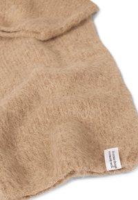 Textile en laine beige avec une texture douce et duveteuse. Présente une petite étiquette dans le coin avec le nom de la marque. Aspect lisse et drapé.