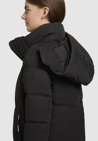 Schwarze Daunenjacke mit hohem Kragen und großer Kapuze. Verfügt über eine Reißverschlusstasche und sichtbare Nähte an den Ärmeln. Weiche, matte Textur.