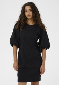 Vestido negro con tejido texturizado, mangas ligeramente abullonadas y una silueta ajustada, que termina justo por encima de la rodilla.