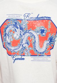 White t-shirt featuring a blue and orange dragon graphic with floral accents. Text reads "Relaxing Spaces," "Henrokuenorem," and "Garden."