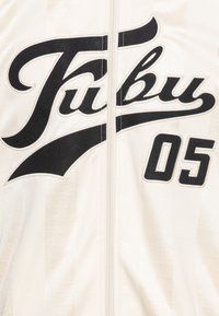 Knitani džemper zip-up u boji off-white s crnim izvezenim "Fubu" logotipom i detaljem "05", s blagim prugama i glatkom teksturom.