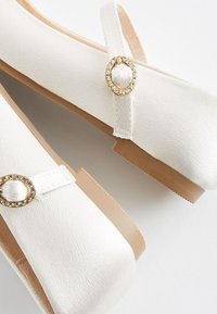 Vita sandlettor i satin har en rund tå, platt sula och en dekorativ accent i guld och rhinsten på remmen.