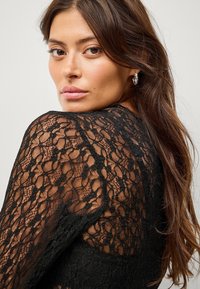 Sort blonde top med intrikate blomster mønstre, en semi-transparent tekstur og lange ærmer. Designet har en rund udskæring og en tætsiddende silhuet.