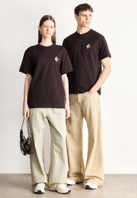 Czarne bawełniane t-shirty z diamentowym logo, zestawione z jasnozielonymi i beżowymi spodniami o szerokich nogawkach. Dodatki obejmują małą czarną torebkę i trampki.