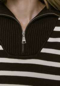 Pull à fermeture éclair rayé marron et blanc avec un col côtelé, présentant une fermeture éclair en métal et une texture douce.