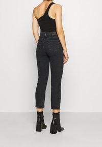 Mujer de pie de espaldas, vistiendo una blusa negra de un solo hombro, jeans negros de talle alto y botas de tacón negro hasta el tobillo.