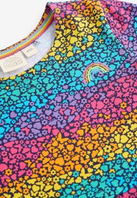 Bunt gemustertes Kleid mit einem Blumen- und Herzmuster sowie Schmetterlingen auf einem Farbverlauf-Hintergrund. Enthält ein Regenbogen-Stickdetail.