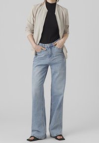 Beige bomberjack, zwarte geribde top, lichtblauwe hoge, uitlopende jeans, gedragen met zwarte instapsandalen. Handen in de zakken, neutrale achtergrond.