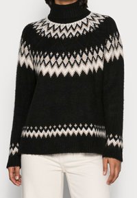 Schwarzer Strickpullover mit hohem Kragen, der ein weißes und beiges geometrisches Muster auf der Brust und den Ärmeln aufweist. Weiche Textur.