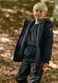 Fant z mornarsko modro jakno z kosmatim podlogom, črno majico in črne hlače z belimi črtami. Naravno okolje z listnim odpadkom.